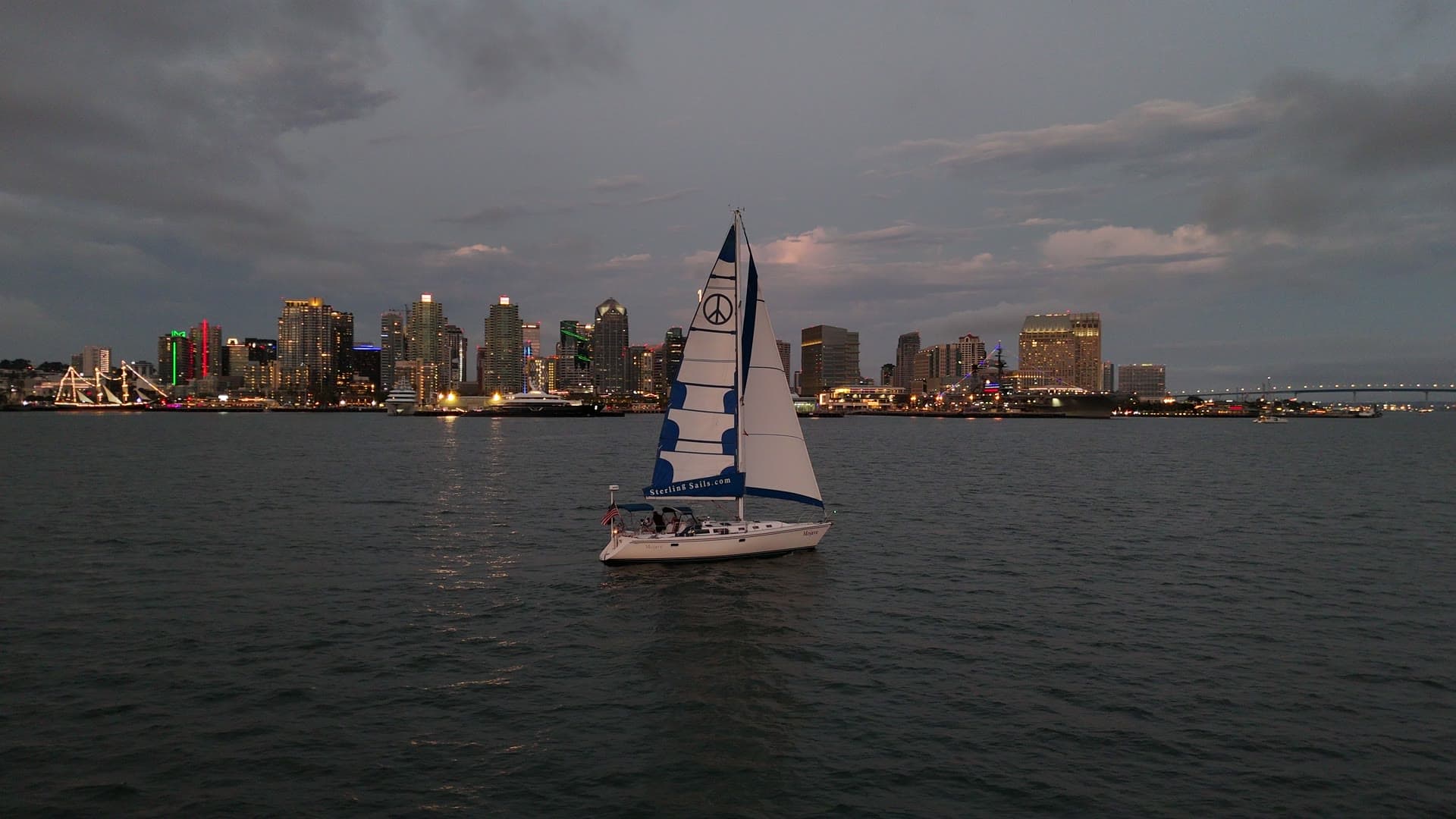 Aerial view of the Mojave under sail past the lit San Diego skyline at dusk